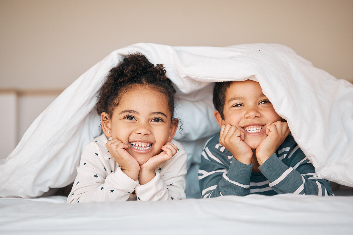 two siblings smiling with their heads peaking out from under a blanket