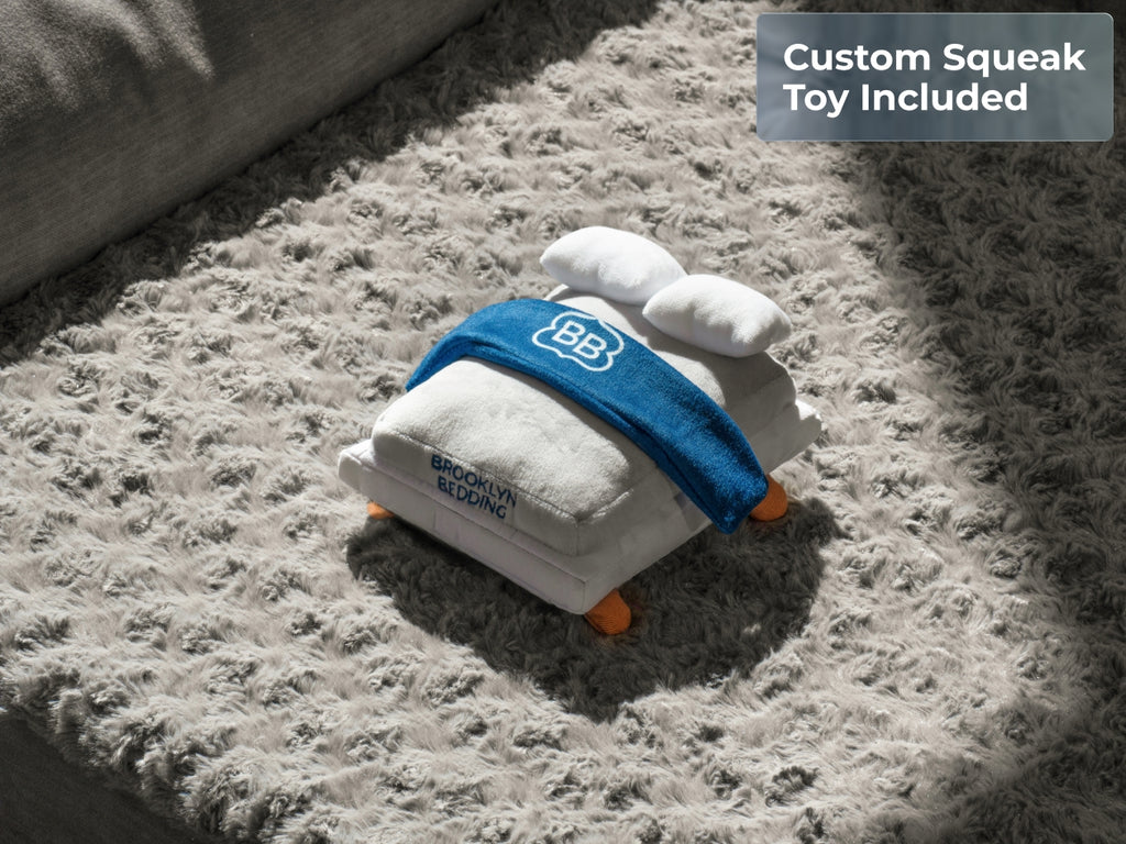 Close-up of a Brooklyn Bedding mattress-shaped squeak toy on the gray textured pet bed. Text reads “Custom Squeak Toy Included.”