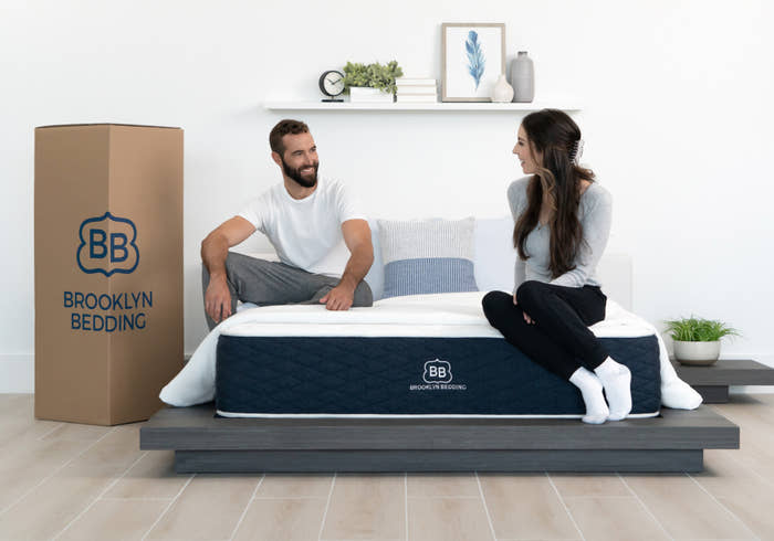 Man and woman sitting on a Brooklyn Bedding mattress in a room with a cardboard box and plants.