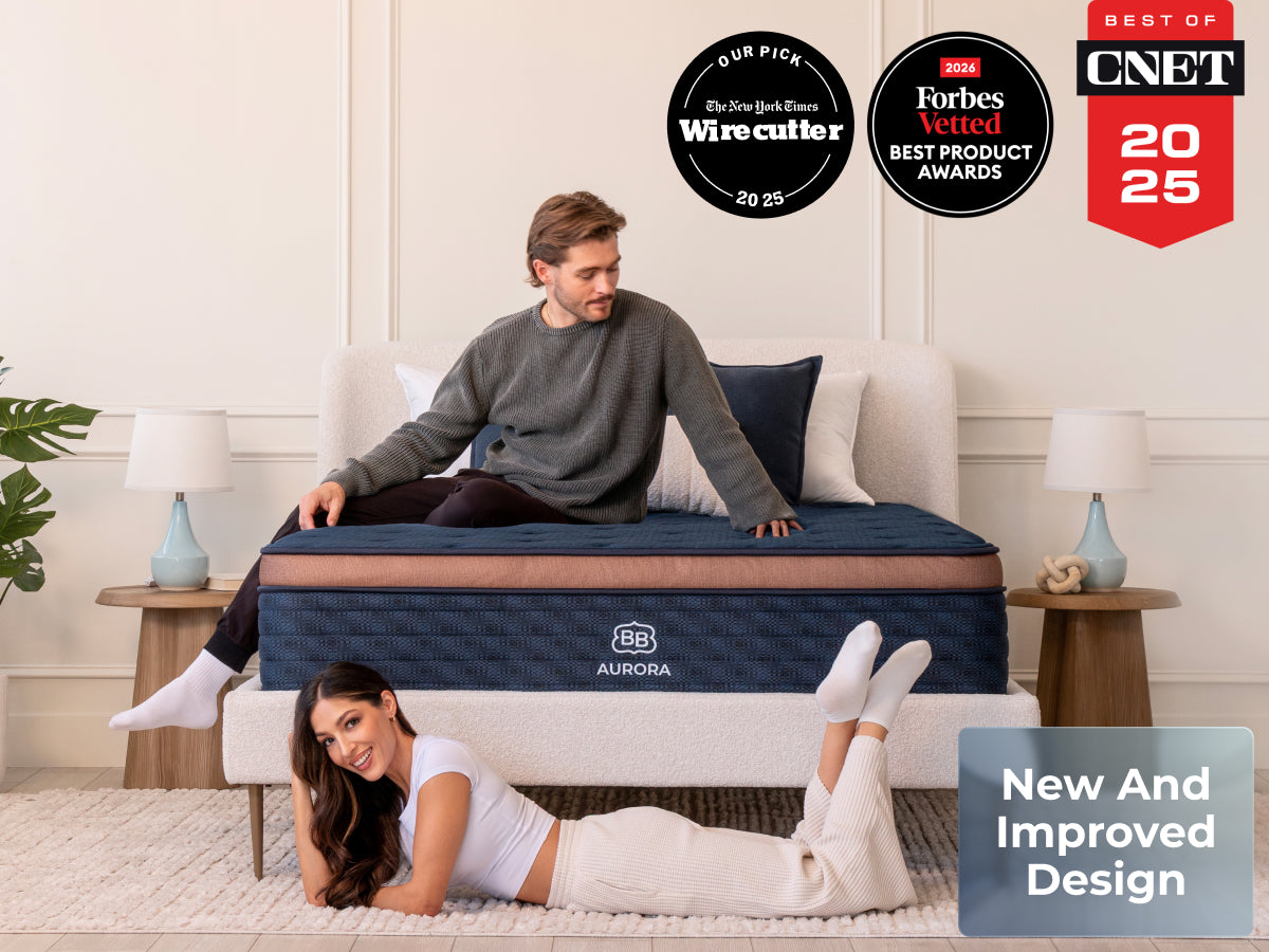 Brooklyn Bedding Aurora Luxe Cooling mattress with two people in a bedroom, with badges reading “Our Pick The New York Times Wirecutter 2025,” “2026 Forbes Vetted Best Product Awards,” “Best of CNET 2025,” and “New And Improved Design.”