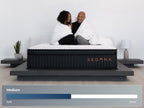 Sedona Elite mattress with two people wrapped in white blankets sitting on the bed, with a Medium firmness scale shown below.