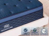 Close-up of the Brooklyn Bedding Signature Hybrid showing the blue quilted top, side panel, and badges reading “Fiberglass Free,” “GREENGUARD Gold,” and “CertiPUR-US.”