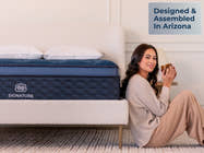 Brooklyn Bedding Signature Hybrid mattress beside a woman sitting on the floor holding a mug, with a “Designed & Assembled In Arizona” callout in a softly lit bedroom.