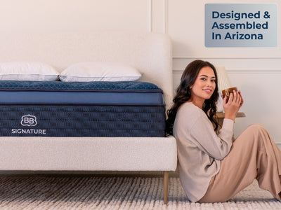 Brooklyn Bedding Signature Hybrid mattress beside a woman sitting on the floor holding a mug, with a “Designed & Assembled In Arizona” callout in a softly lit bedroom.