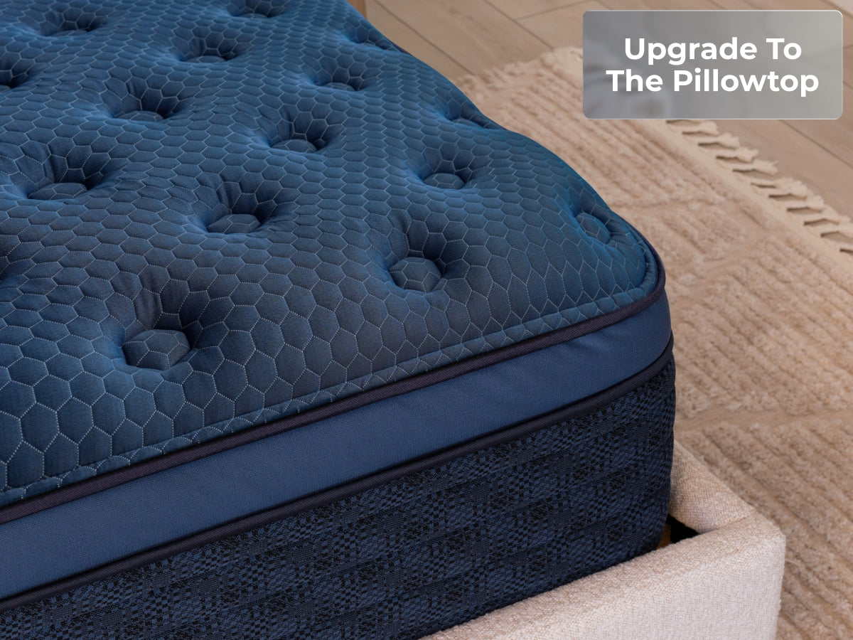 Close-up of the Brooklyn Bedding Signature Hybrid pillow top showing the tufted blue surface and side panel, with a badge reading “Upgrade To The Pillowtop.”