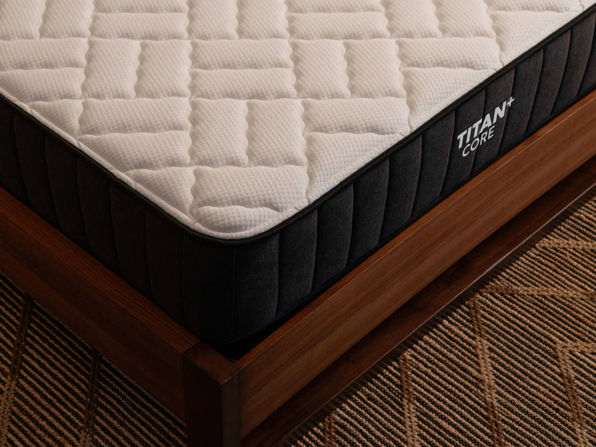 Close-up corner view of the Brooklyn Bedding Titan Plus Core mattress resting on a walnut wood bed frame, showing the cream-colored quilted top panel and charcoal ribbed border.