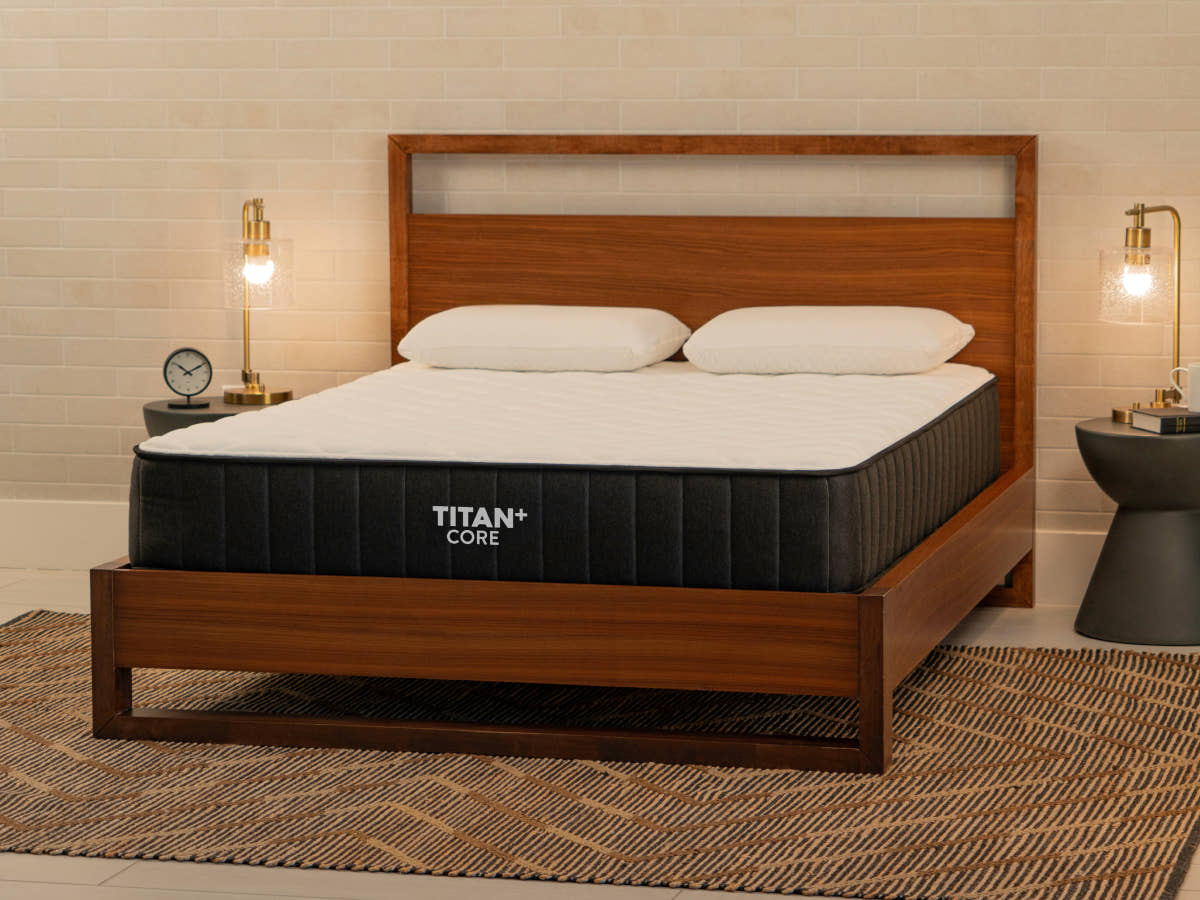  Brooklyn Bedding Titan Plus Core mattress on a walnut wood platform bed in a warm, mid-century modern bedroom with two white pillows, brass sconce lamps, and a woven area rug.