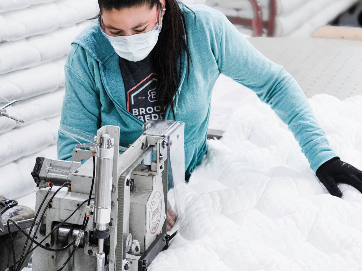 A person wearing a mask operates a sewing machine, stitching white quilted fabric in a textile or bedding manufacturing setting.