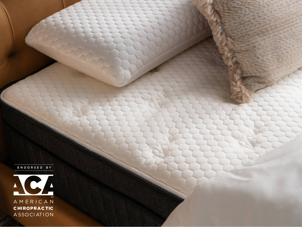 A close-up view of a Titan Plus Luxe with a hexagonal patterned mattress cover, a white pillow, and a textured decorative pillow. The image includes a logo for the American Chiropractic Association.