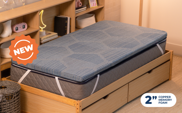 Person sitting cross-legged on the CopperFlex 201 Mattress Topper on a bed with headboard shelving and string lights.