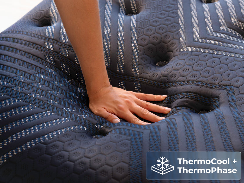 Hand pressing down into the quilted, textured top of the navy blue mattress. Text overlay: "ThermoCool + ThermoPhase" next to a snowflake icon.