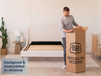 Man opening a tall Brooklyn Bedding box next to an empty bed frame. Text overlay: "Designed & Assembled In Arizona."