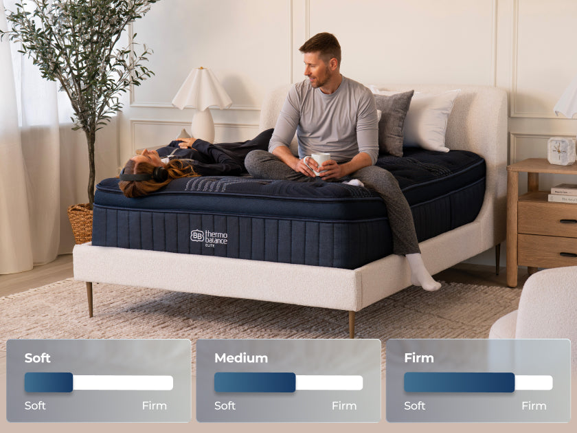 Woman lying down with headphones while a man sits beside her on the mattress. Graphic overlay displays slider scales for "Soft," "Medium," and "Firm."