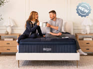 Smiling couple sitting on a navy blue ThermoBalance Elite mattress holding coffee mugs. Text overlay: "NEW" badge.