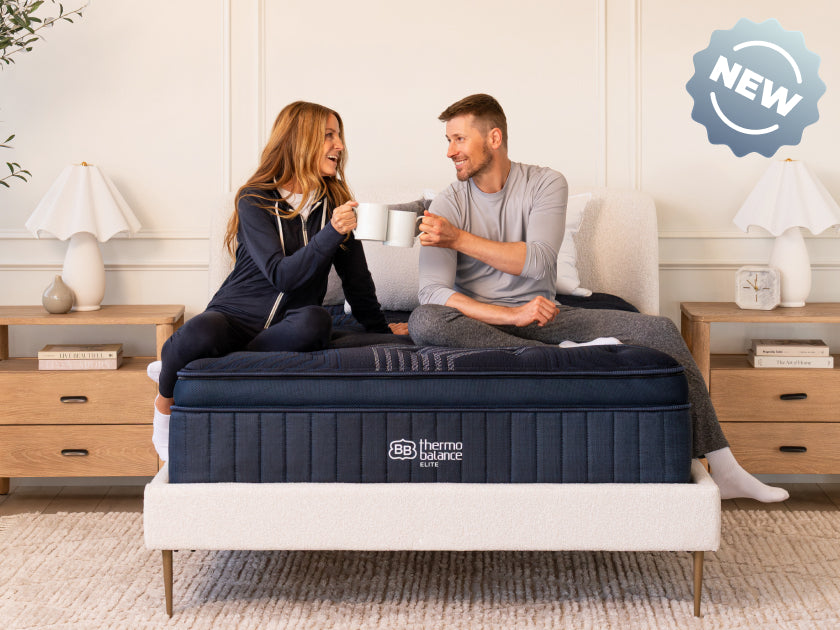 Smiling couple sitting on a navy blue ThermoBalance Elite mattress holding coffee mugs. Text overlay: "NEW" badge.