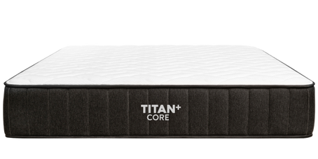 Brooklyn Bedding Titan Plus Core mattress on a rust-colored upholstered bed frame in a stylized bedroom setting with two people relaxing on top. Badge reads: 